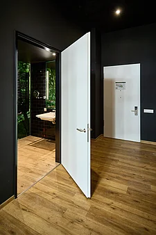 Innenansicht eines Zimmers mit hellem Holzfußboden und dunklen Wänden. Im Vordergrund eine geöffnete weiße Innentür in schwarzer Zarge mit silbernem Türgriff, die in ein Bad mit schwarzer Fliesenwand, Waschbecken und beleuchtetem Spiegel führt. Im Hintergrund eine geschlossene weiße Wohnungstür mit Schloss, Türspion und Informationsblatt.