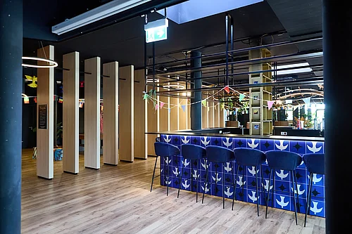 Offener Innenraum eines Campus: blau gekachelte Bar mit mehreren hohen blauen Hockern, heller Holzfußboden, vertikale Holzlamellen als Raumtrenner, hängende Lichtringe, bunte Wimpelgirlanden, Pflanzen und dunkle Decke mit sichtbarer Haustechnik.