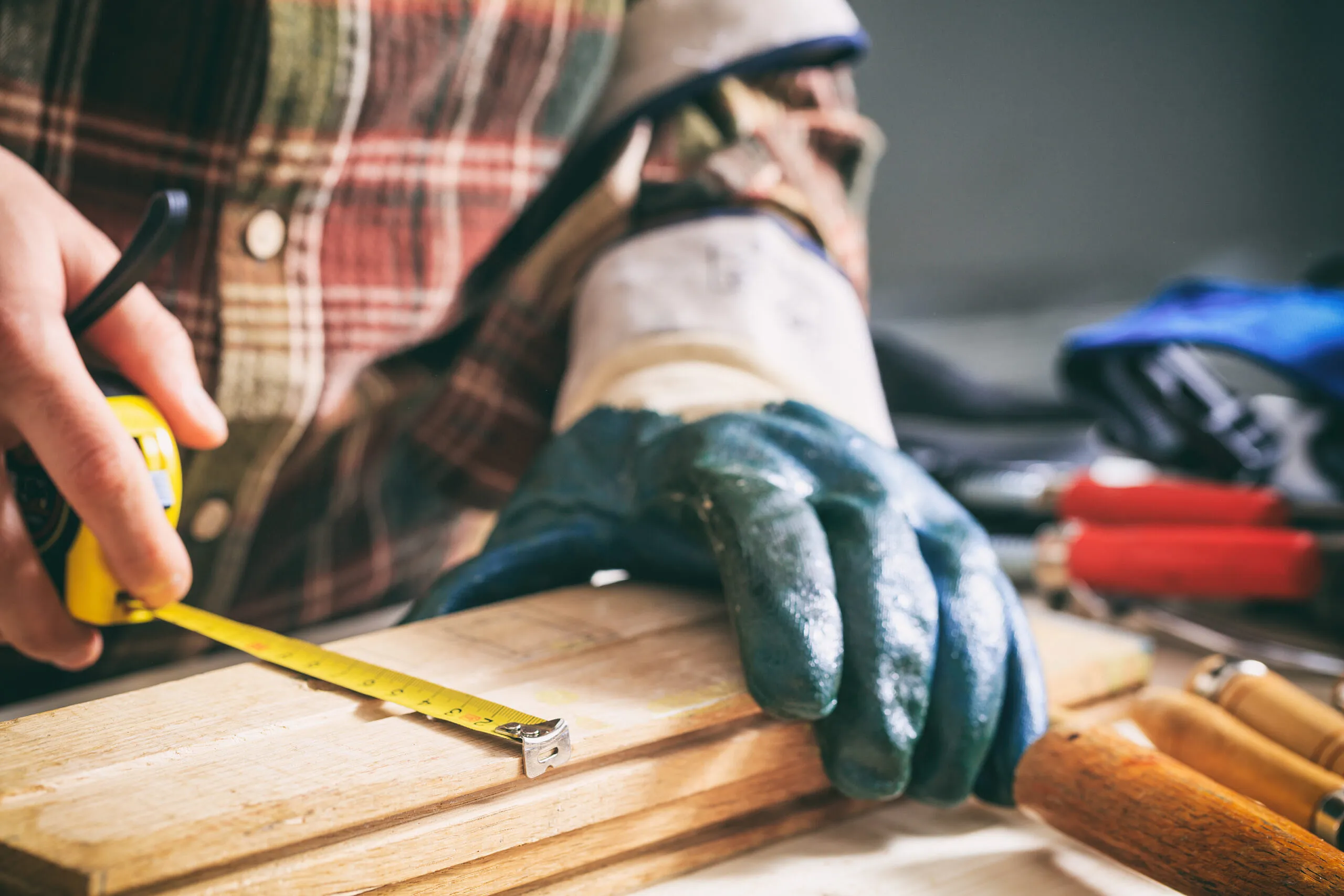 Hand zieht ein gelbes Maßband über ein Holzbrett, andere Hand stabilisiert das Brett mit blauem Arbeitshandschuh; unscharfe Werkzeuge und kariertes Hemd im Hintergrund