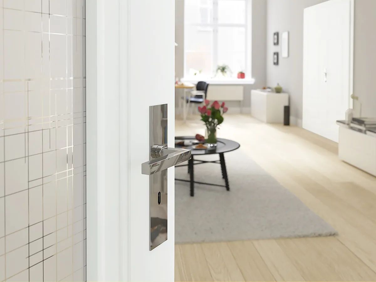 Weiß lackierte Innentür teilweise geöffnet, glänzender rechteckiger Edelstahl-Türgriff mit Langschild und Schlüsselloch im Vordergrund; im Hintergrund helles Zimmer mit Parkettboden, grauem Teppich, rundem schwarzen Couchtisch mit Vase und roten Tulpen, Fenster mit Tageslicht, Schreibtisch und Stuhl